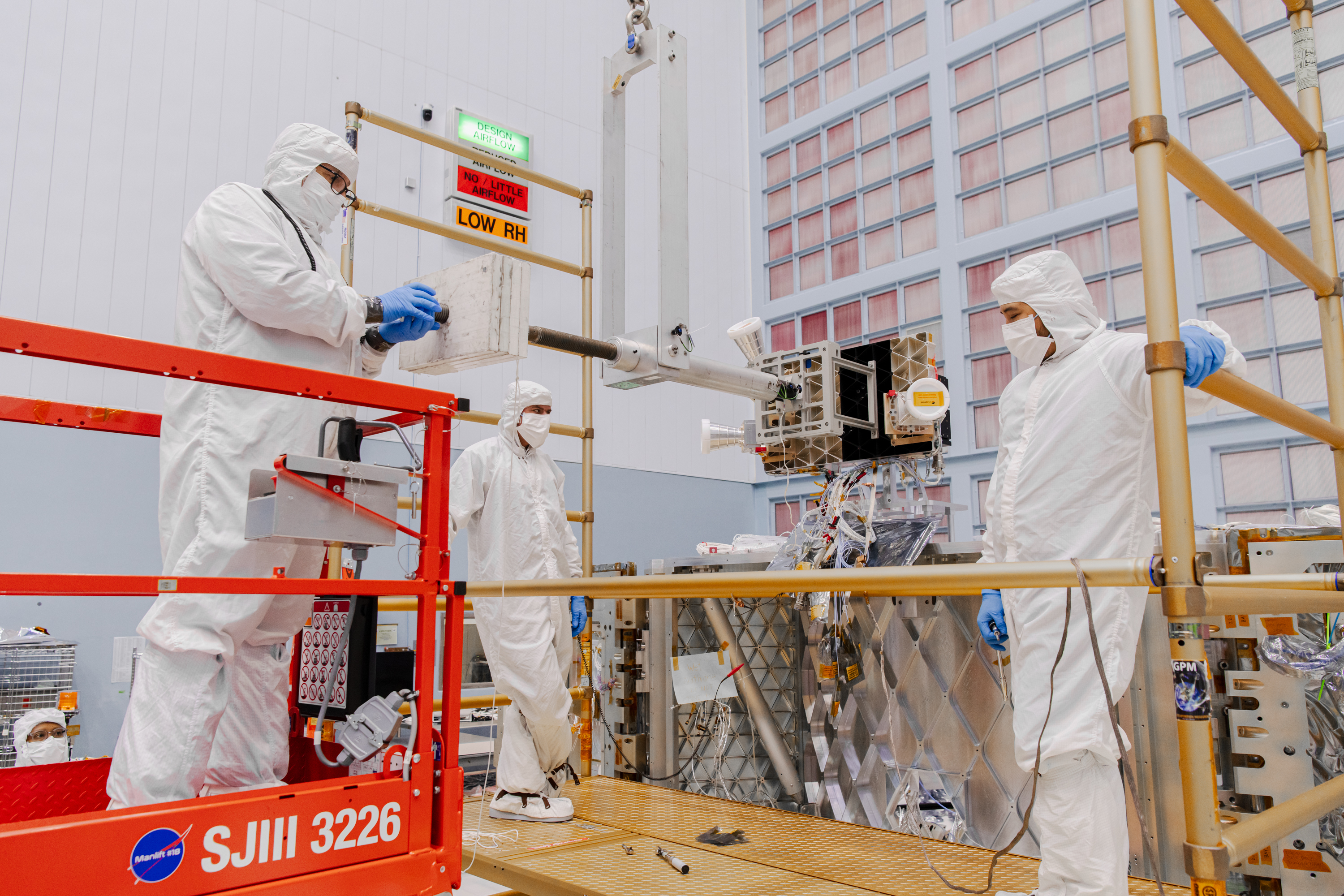 People are in head-to-toe white clean room jumpsuits with blue gloves. They are standing on a gold-colored platform facing forward. In the foreground is a device suspended horizontally from hanging metal rods. At the end of the rods is a large square box, with three white horn-shaped instruments around it. The back wall is covered in a grid of light pink squares, which are air filters. Photo credit: NASA/ Sydney Rohde