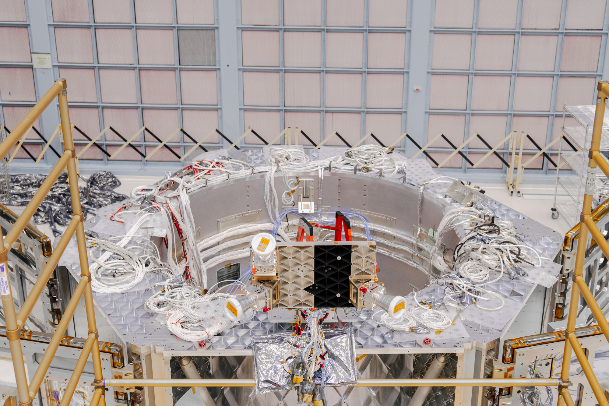 Top-down view of a large silver hexagonal-shaped structure with a circular opening inside. Tons of white wires are bunched on top of the structure. In the foreground is a large square box with three white horn-shaped instruments around it. The back wall is covered in a grid of light pink squares, which are air filters. Photo credit: NASA/ Sydney Rohde