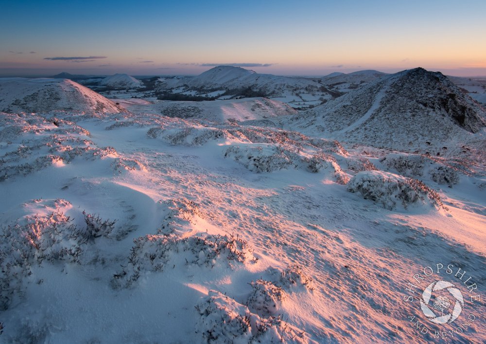 🎄Day 6 of our #Shropshire advent calendar takes us to the Long Mynd, with sunrise casting a delicate pink light across the snow-covered landscape. The Iron Age hill fort of Bodbury Ring is on the left, with Burway Hill to the right and Caer Caradoc on the horizon. 🎅