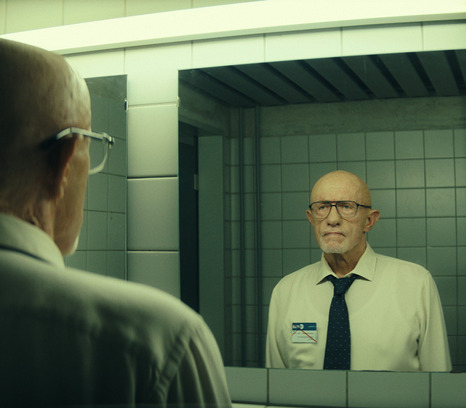 From Constellation, Jonathan Banks stands in a bathroom with white tiled walls, looking into a mirror at himself. He wears a white button-down shirt, black tie, black rectangular eye-glasses, and a name tag. He has a serious expression on his face.