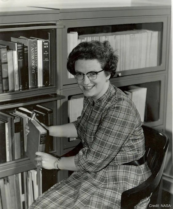 In this undated image from the 1960s, Nancy Grace Roman sits in her office in NASA Headquarters. Dr. Roman has dark, curly hair and wears black, horn-rimmed glasses. She is wearing a plaid dress with elbow-length sleeves. She sits in front of bookcases with glass-paneled doors, some of which are open as she removes a book. A few of the book titles are legible, including “Statistical Astronomy,” “The Sun,” and “Stellar Structure.” The image is watermarked “Credit: NASA.”