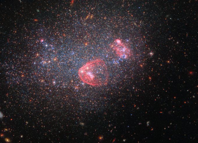 A collection of stars and galaxies fill the scene against a dark background. The image is dominated by a dense collection of stars that make up the irregular galaxy UGC 8091. The stars span a variety of colours, including blue and orange, with patches of blue occupying the central part of the galaxy. There are also visible circular regions of red/pink gas within the galaxy.