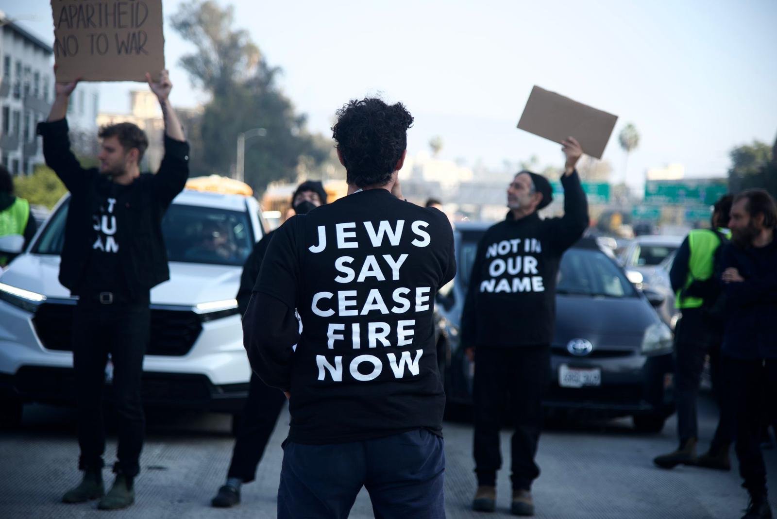 3 people are standing, blocking a line of traffic. They are wearing black t-shirts that say "Jews say ceasefire now" in white writing on the back, and "not in our name" on the front. In the background you can see skyscrapers, freeway signs, and a long line of cars.