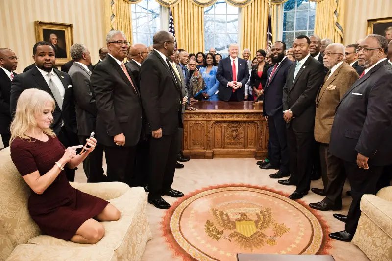 Counselor to the President Kellyanne Conway checks her phone as President Donald Trump and leaders of historically black universities and colleges pose for a group photo in the Oval Office(2017)