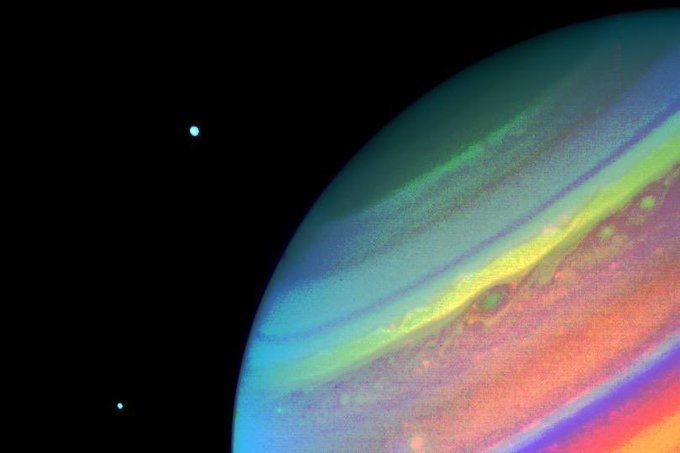 In this Voyager 2 image of Saturn, obtained Aug. 11, 1981, from a range of 14.7 million kilometers (9.1 million miles), north is at the upper right edge of the disc. Seen above the planet are the satellites Dione (right) and Enceladus. This false-color print shows a green spot at the south edge of a yellow band; in true color, the spot would appear brown and the band white. A bright yellow spot slightly above and to the left in this image moves eastward relative to the green spot at a rate that allows it to pass the green feature in about 50 days. The convective clouds that appear between the two spots are typical of the region. Caption credit: NASA