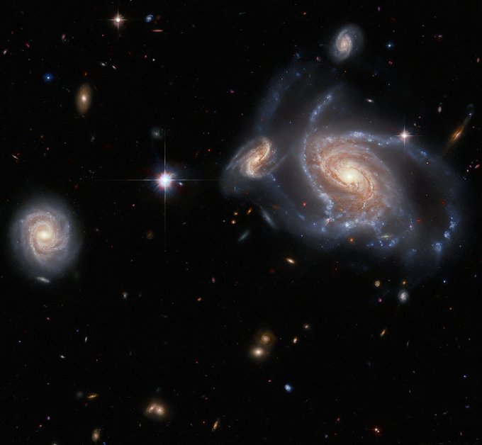 A collection of galaxies. On the left side a large spiral galaxy with swirling, twisted arms is flanked by a smaller, but still detailed, spiral behind its arm on the left, and a smaller spiral above it. On the right side is a fourth, round spiral galaxy seen face-on. Between them lies a single bright star. Several stars and distant galaxies dot the background.