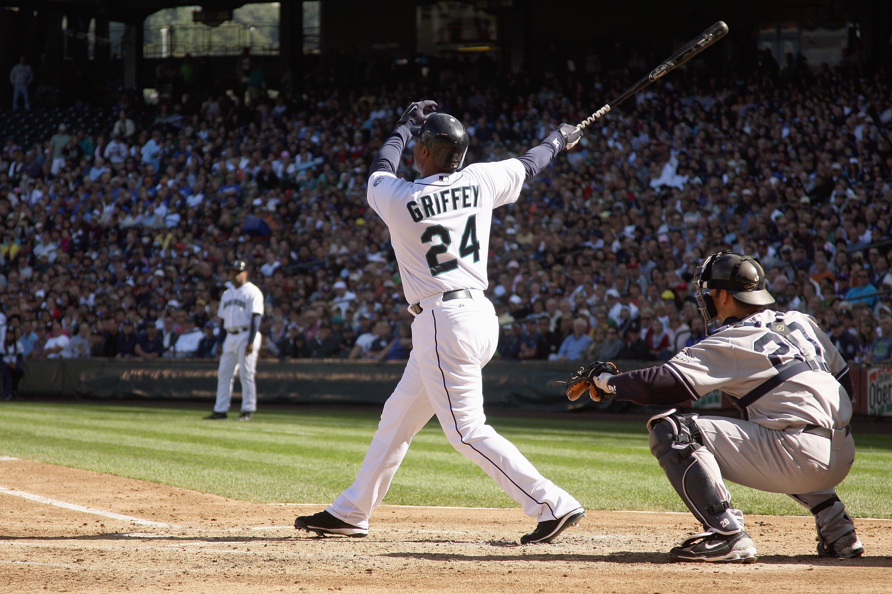 Ken Griffey Jr. follows through on a swing. He is wearing a white Seattle Mariners jersey with “Griffey” and “24” in black and northwest green lettering on the back. 