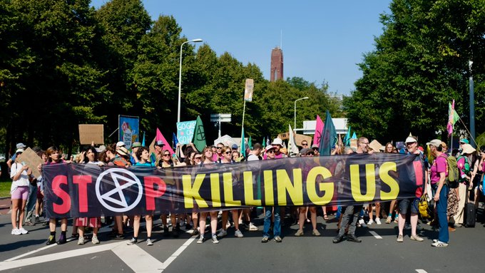 Een grote groep actievoerders met een groot spandoek met de tekst STOP KILLING US, waarbij de O van stop het XR logo is.