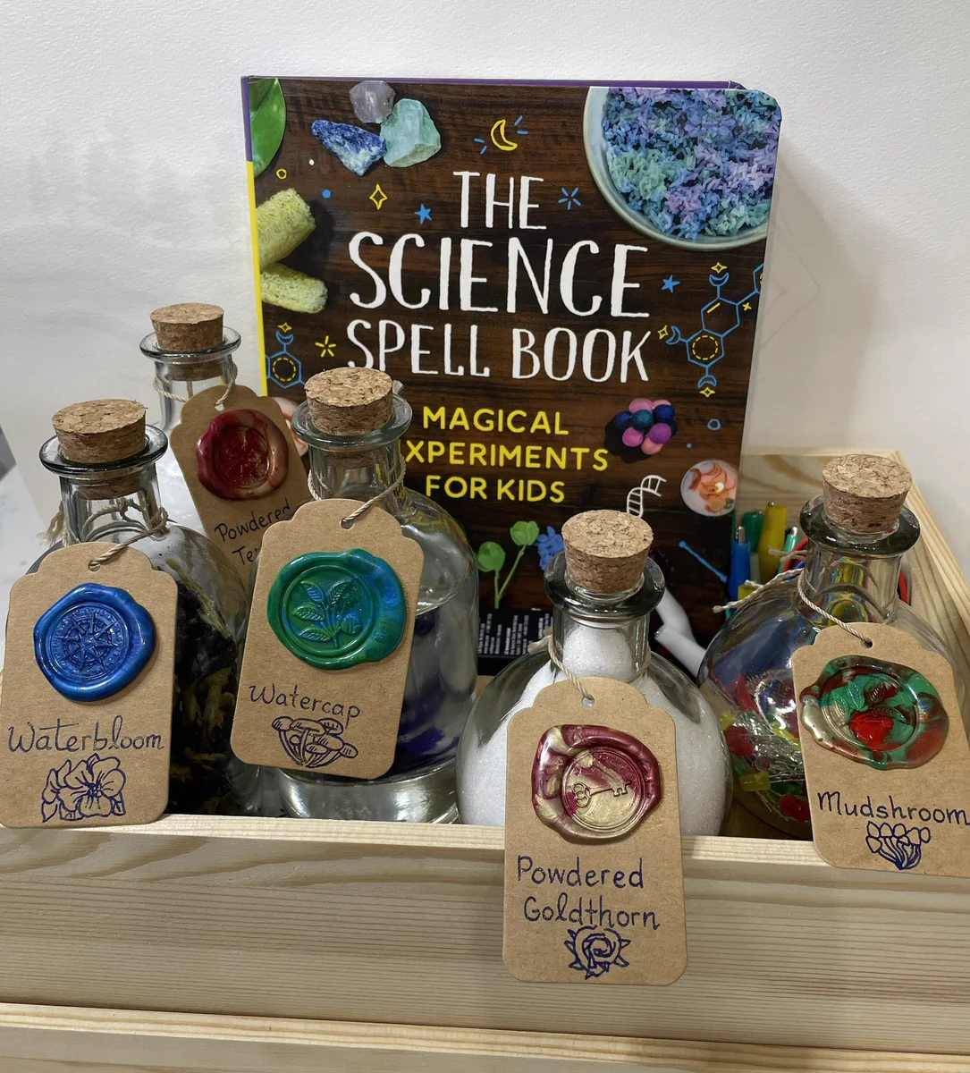Wooden box with potion bottles labeled with wax seals and a book called The Science Spell Book: Magical Experiments for Kids. The bottles are labeled Waterbloom, Watercap, Powdered Goldthorn, Mudshroom, and in the back Powdered Terraria.