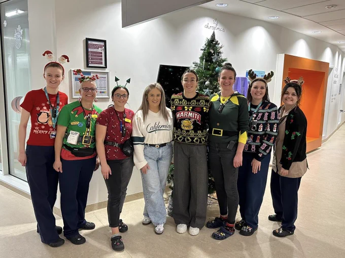 Eight females stand in festive attire in front of a Christmas tree on a hospital ward.