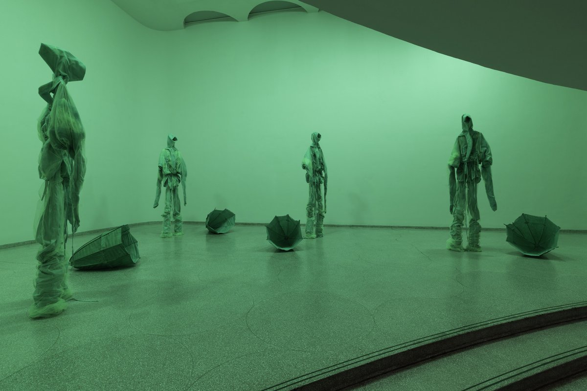 Four figures, approximately 9 feet tall, loom large in a room saturated with an acid-green light. They are heavily concealed and seem mysterious. Hoodies shield “heads” that are completely hidden from view. Even as these beings have a great presence, they are never wholly