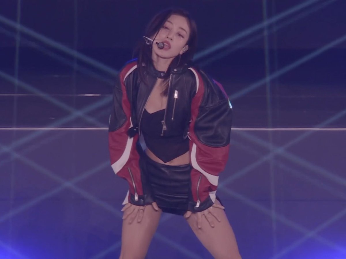 A female performer on stage wearing a black bodysuit, black leather skirt, and a red, black, and white leather jacket, performing under blue stage lighting with laser beams.