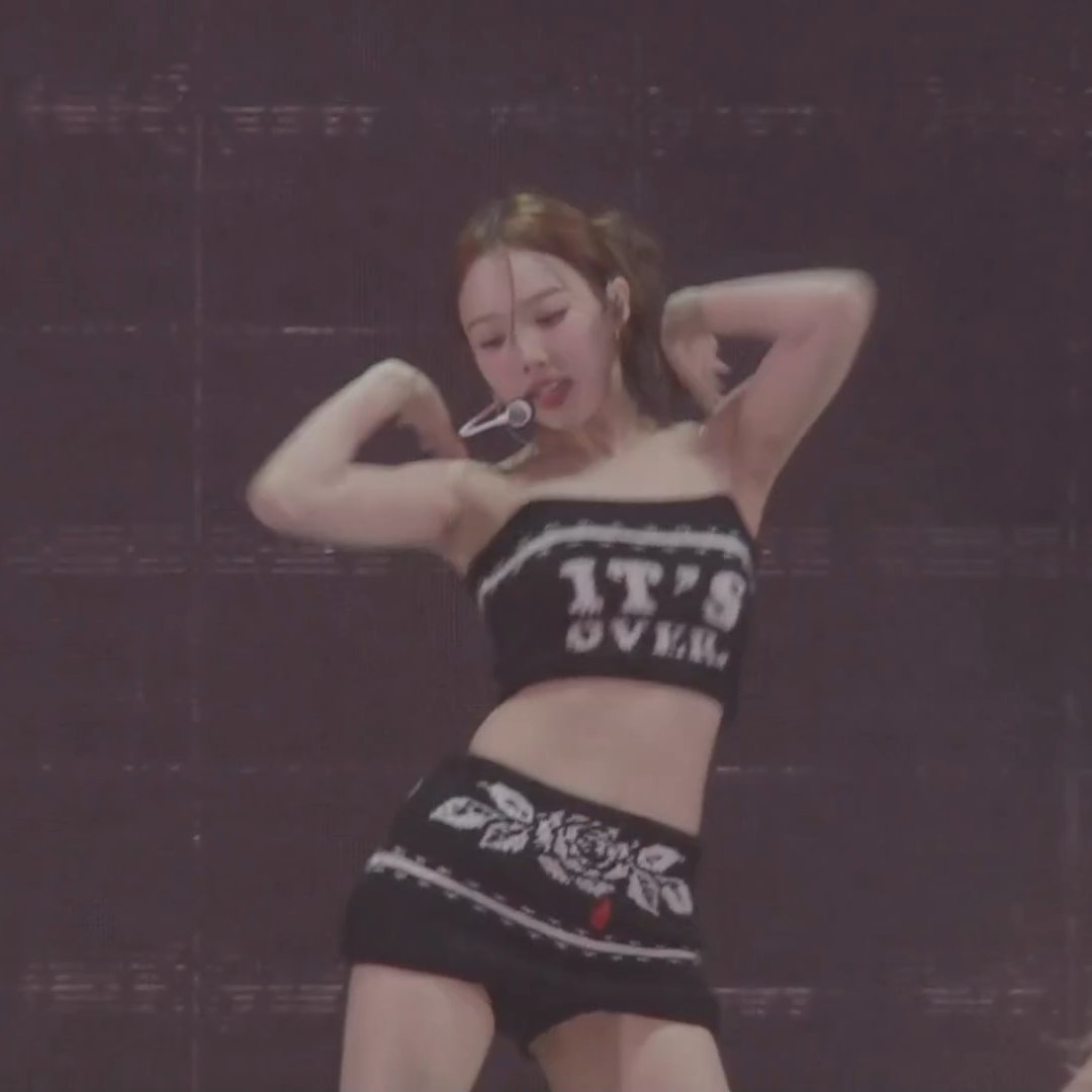 A performer on stage wearing a black tube top reading 'IT'S OVER' and a black mini skirt with a white rose design, posing with both hands behind her head while wearing an earpiece microphone.