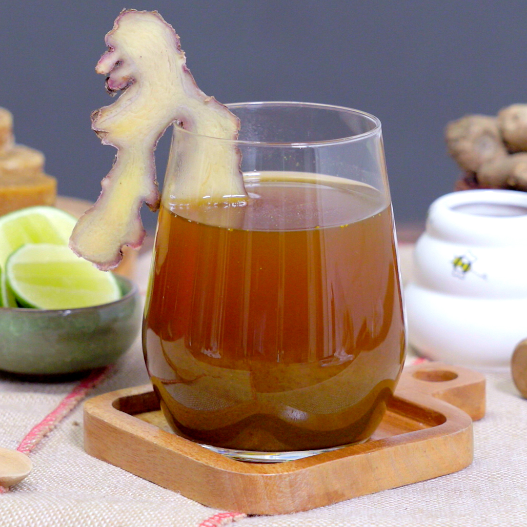 Close-up photo of a glass of amber-colored ginger tea with a large piece of crystallized ginger as garnish, served on a wooden tray with sliced apples and fresh ginger visible in the background.