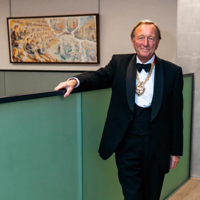 Sir Neil Mortensen stands for a photo wearing a black suit and tie and a with a medal around his neck.