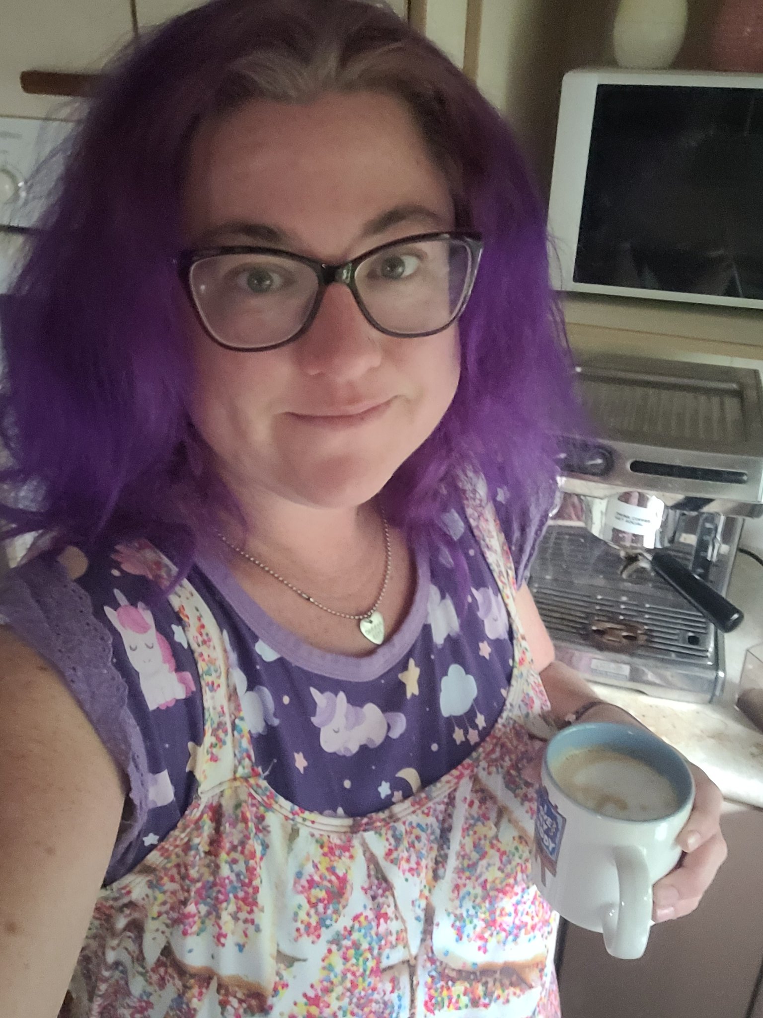 A woman with fluffy purple hair and glasses wearing a purple onesie with lace shoulders and unicorns. She has a romper with fairy bread print over the top. She is holding a coffee in front of a coffee machine
