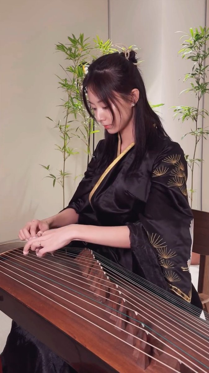 A young woman with long dark hair, wearing a black traditional garment with gold trim and pine tree embroidery, plays a guzheng (a Chinese stringed instrument). Bamboo plants are visible in the background.