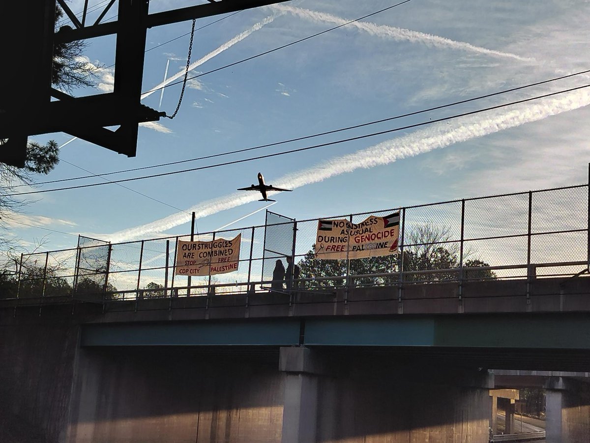 Two banners hanging on a bridge with an airplane flying behind it. “NO BUSINESS AS USUAL DURING A GENOCIDE”