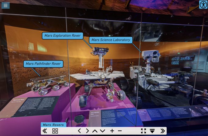 Three rovers on display in a museum exhibition case. A small rover labeled Mars Pathfinder Rover, a mid-sized rover labeled Mars Exploration Rover and a car-size rover labeled Mars Science Laboratory.
