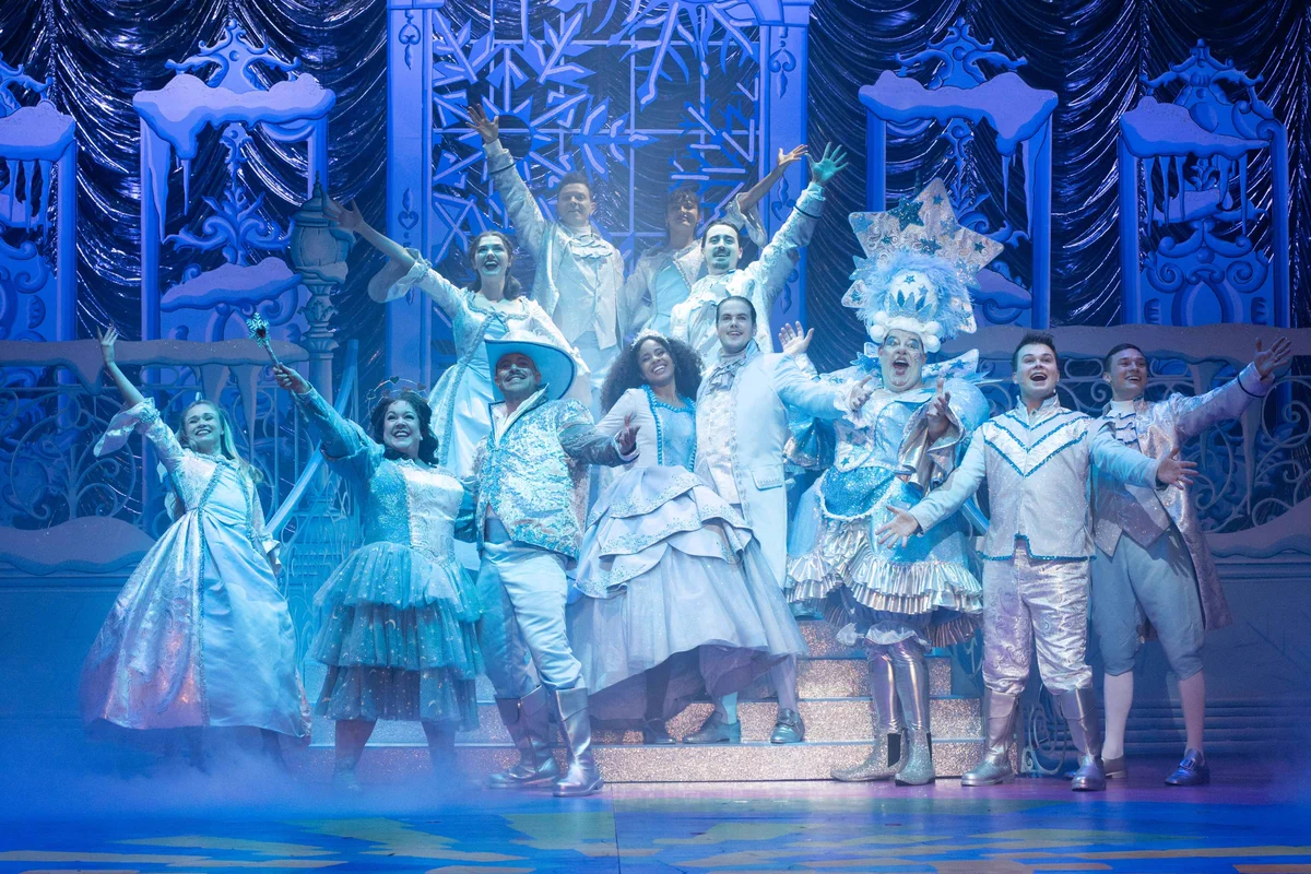 Production image with the full company of Beauty and the Beast posing with their arms open wide during the walkdown and bows. They wear light blue and white costumes and stand in front of a frost backdrop with snowflakes.
