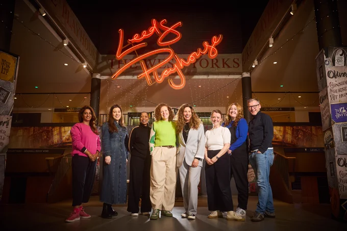 Eight people stood in a line. They are under a neon banner saying Leeds Playhouse