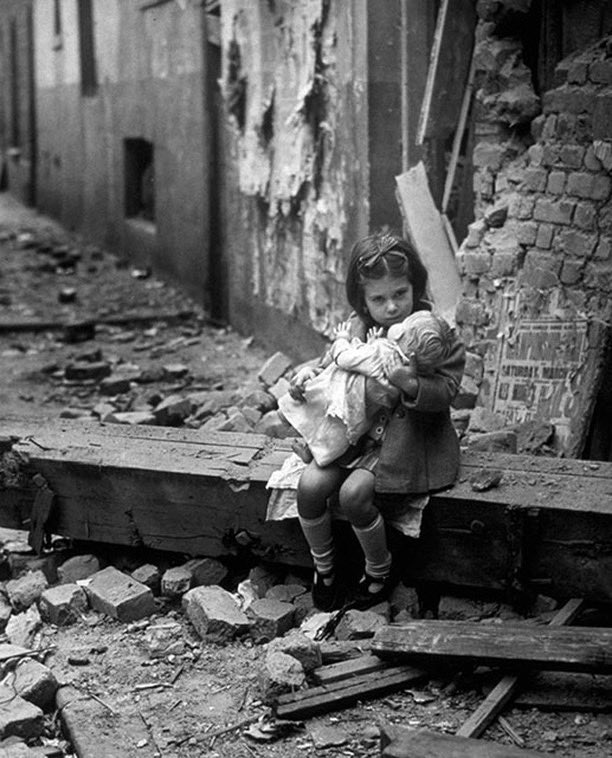 In 1940 London, a young girl sits among the debris of her bombed house, holding her doll.