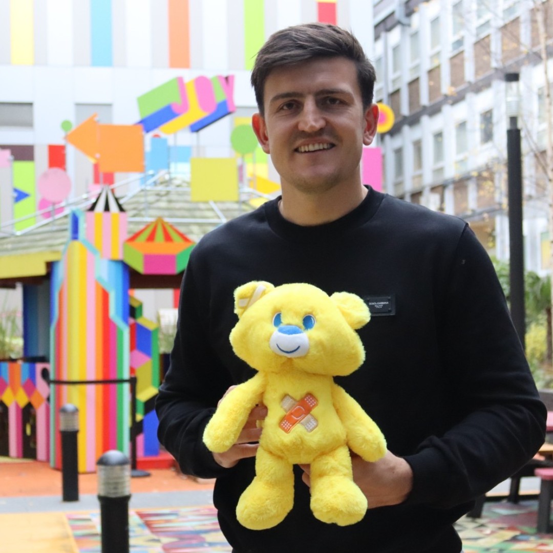 Footballer Harry Maguire stands smiling with a small yellow teddy bear in front of a bright multi-coloured building.