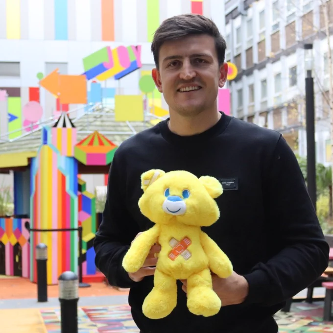 Footballer Harry Maguire stands smiling with a small yellow teddy bear in front of a bright multi-coloured building.