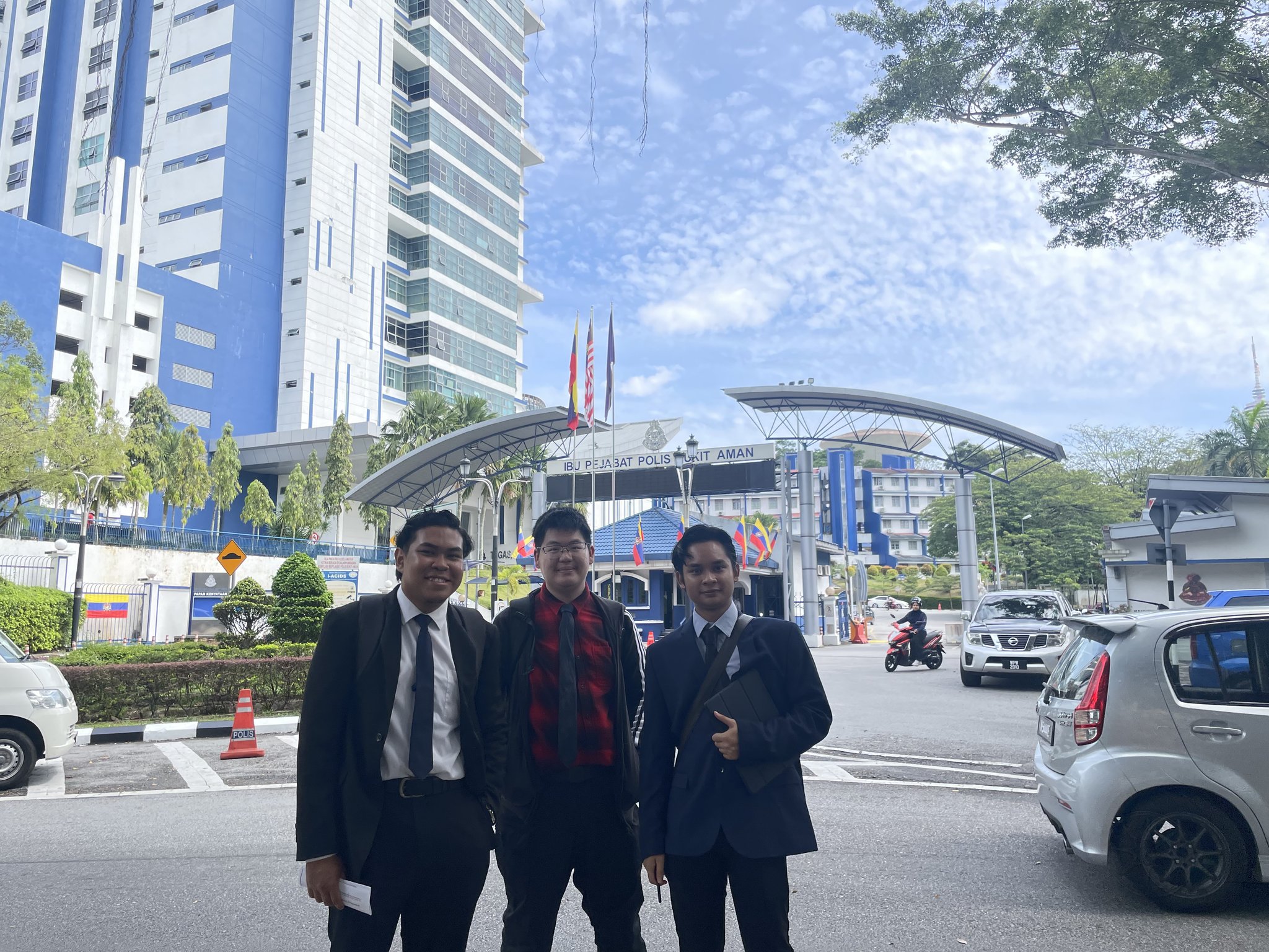 3 young adult males stand in front of the entrance to a police headquarters building.