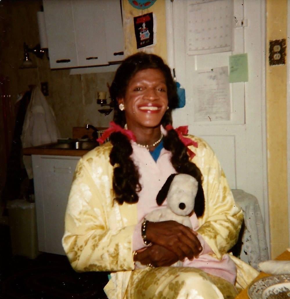 A rare photo captures Marsha P. Johnson wearing ponytails adorned with bow ties, radiating adorable charm and a beautiful smile. Clutching a Snoopy teddy bear, it reflects Marsha's affection for bears and dolls. The date is unknown, and credit goes to the photographer. 📸