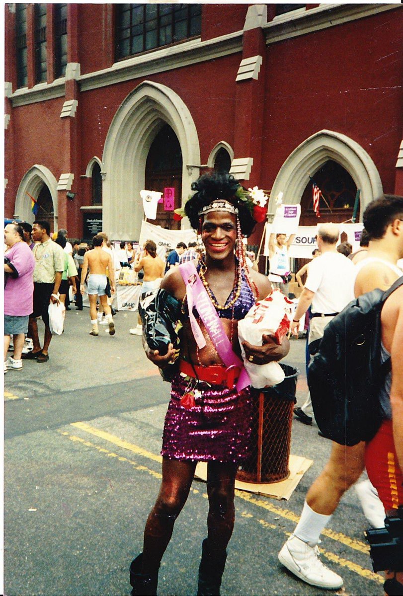 A rare photo captures Marsha P. Johnson at an event, donning a dazzling blue bra paired with a pink sparkly skirt adorned with jewelry. Her iconic look includes stunning hair, adorned with flowers, and, of course, her radiant smile with a flawlessly beat face. Credit to 📸