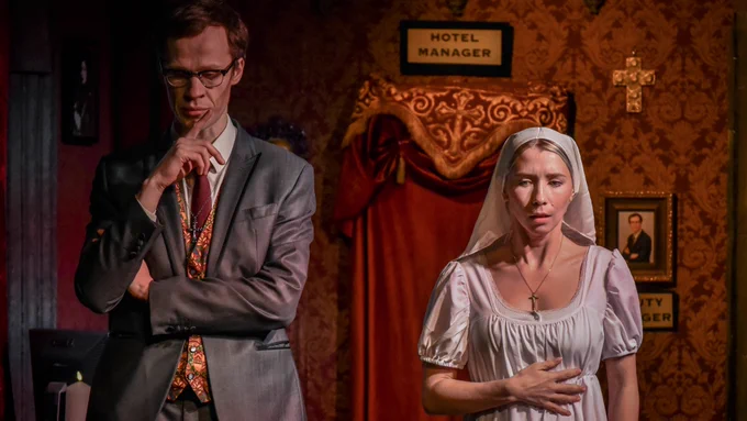 Two actors stand side-by-side, the tension palpable. One wears a grey dark suit, and looks down at the floor, his fingers pressed to his lips. The other, wearing a white dress and nun's habit, looks apprehensive, and clutches her hand to her waist.