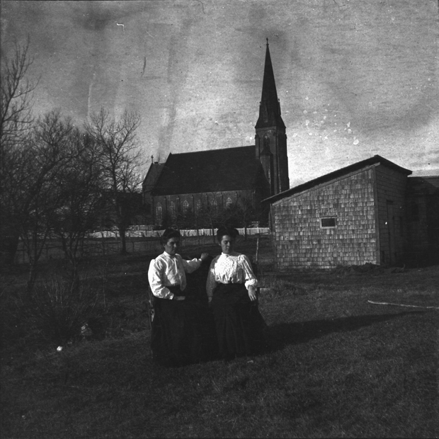 Two women sitting on a bench, St. Simon and St. Jude Church in background, Tignish [ca. 1900] Acc2330/H-50