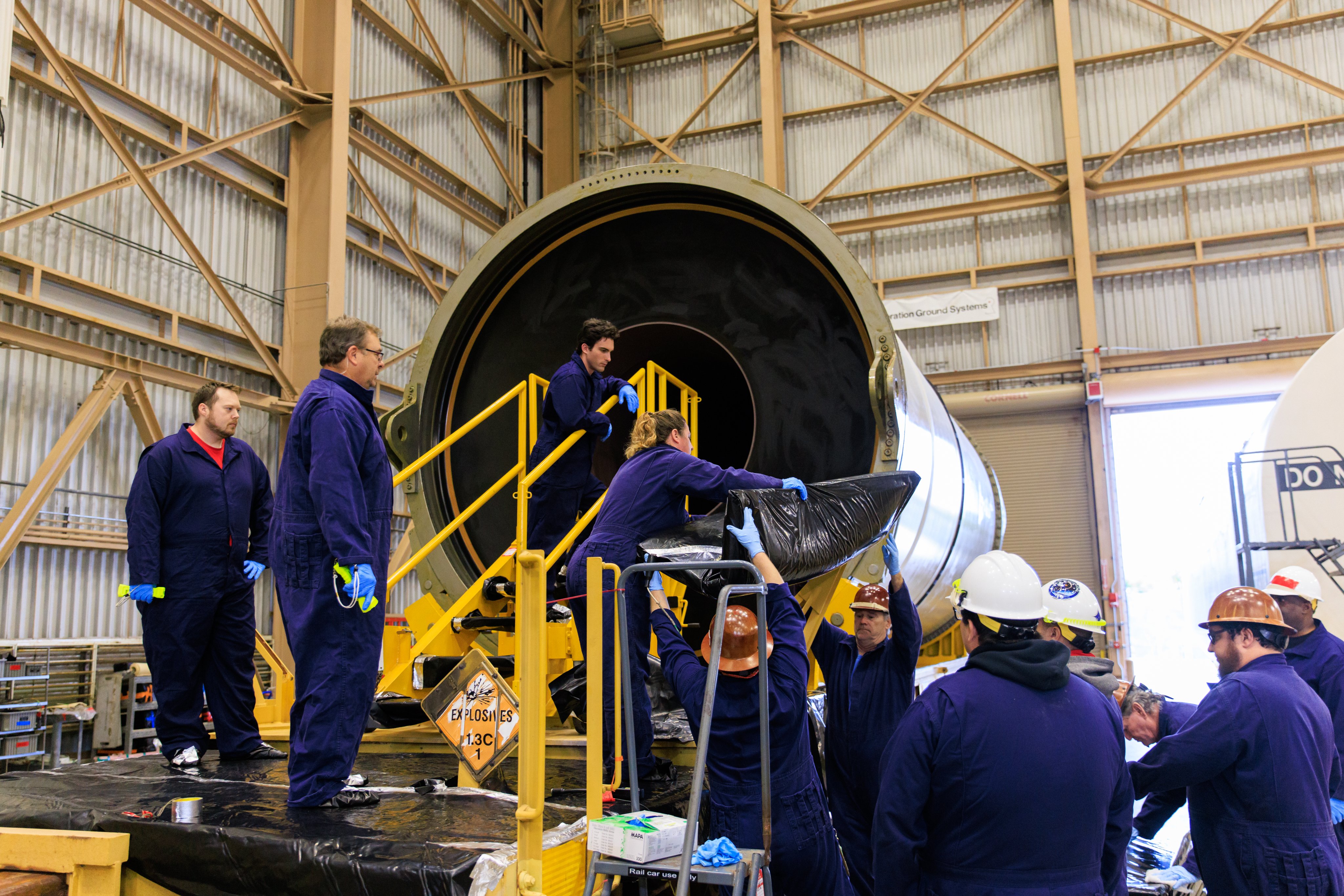 Technicians conduct an inspection of the middle of a horizontal rocket booster segment. A white segment is seen in a large metal building. Photo Credit: (NASA/Isaac Watson)