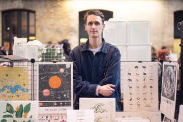 Illustrator Alex Foster, a slim guy wearing a blue collared shirt, stands at a table display with a bunch of his posters (one about animals, one about the moon, one with birds of paradise flowers). The Solar System poster featuring Zoozve is one of the posters.
