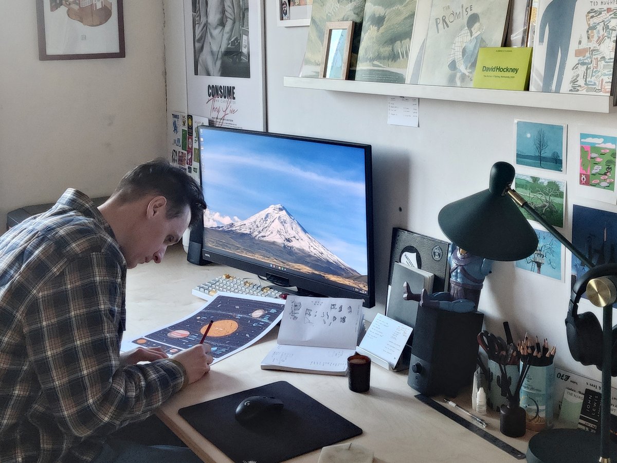 Illustrator Alex Foster sitting at his desk working on the Solar system poster with Zoozve in it. On the desk is a computer, speaker, pencils, lamp, headphones, and so on. Lot of postcards and posters up on the wall of naturescapes and so on.