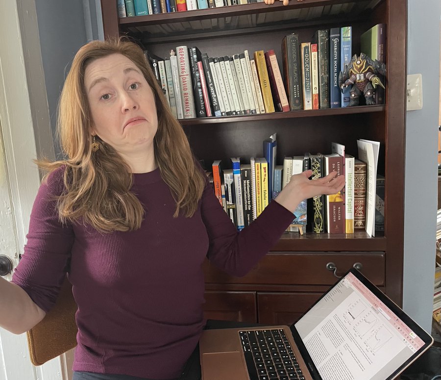 Liz, a woman with long hair and a maroon shirt. Sits in front of a bookcase, at a table with a laptop on it, a scientific paper open on the laptop. She is shrugging. 