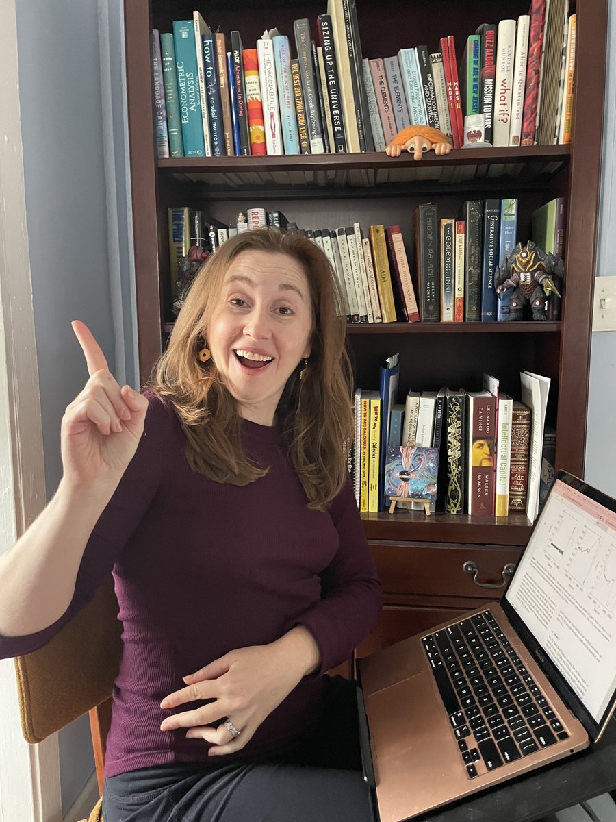 Liz, a woman with long hair, earrings and a maroon shirt sitting in front of a bookcase, at a table with a laptop, holding her finger up like she figured something out!