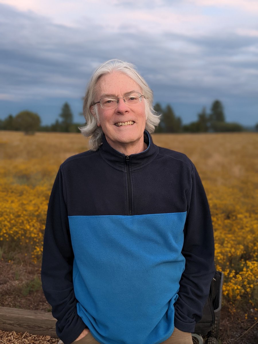 Picture of a smiling Brian Skiff, long silver hair, wearing glasses and a fleece sweater, outside against a background of yellow flowers and a slightly cloudy sky.