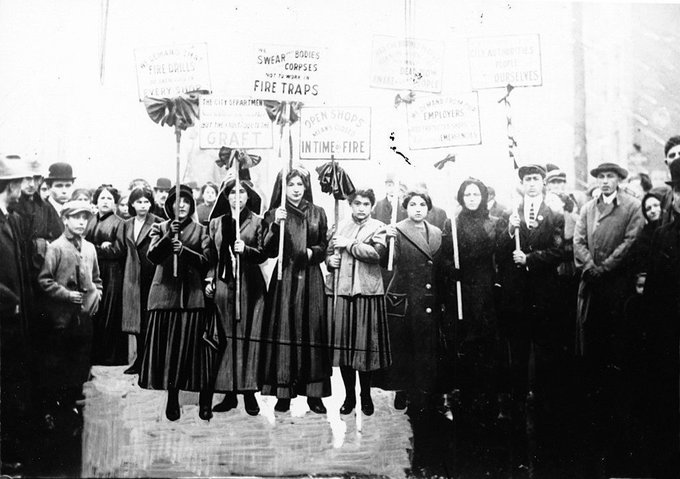 Women wearing black dresses, holding signs that say things about fire drills and fire traps. 