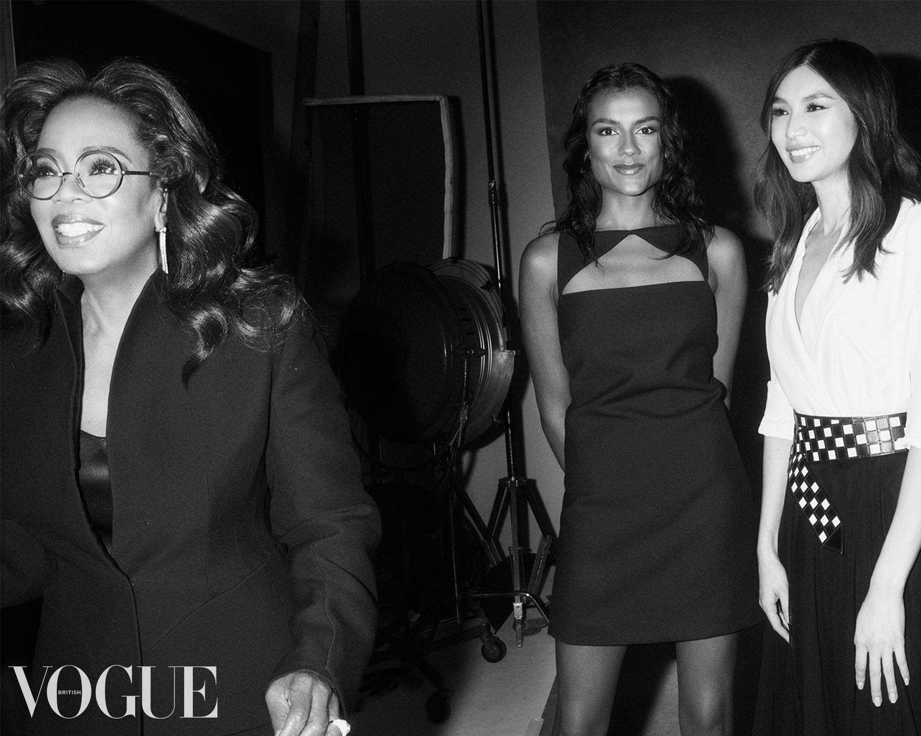 A black and white image shows Oprah Winfrey, a Black woman, visible in the foreground at the left of the frame. She is wearing black tailoring and smiling at something off camera. Behind her stands Simone Ashley, a woman of Indian Tamil descent, who is wearing a black minidress and looking into the camera, and to her left, Gemma Chan, a woman of Chinese descent, who is wearing a white shirt.