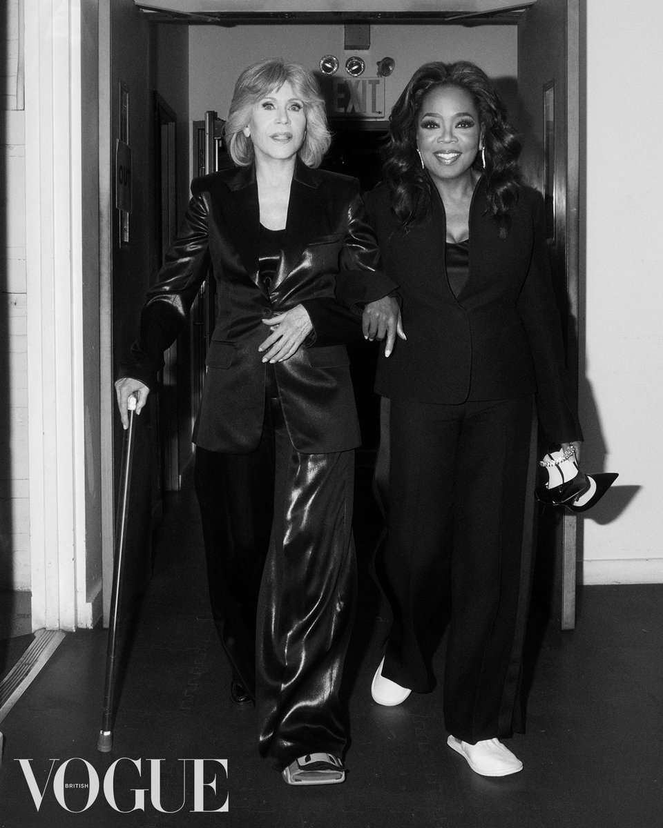 Black and white image shows Jane Fonda, a white woman, and Oprah Winfrey, a Black woman, walking arm in arm down a corridor in a studio space. Jane is wearing a leather suit and her left foot is in a cast. Her right hand rests on a cane. Oprah is wearing black tailoring and white trainers. She is looking directly into the camera and smiling.