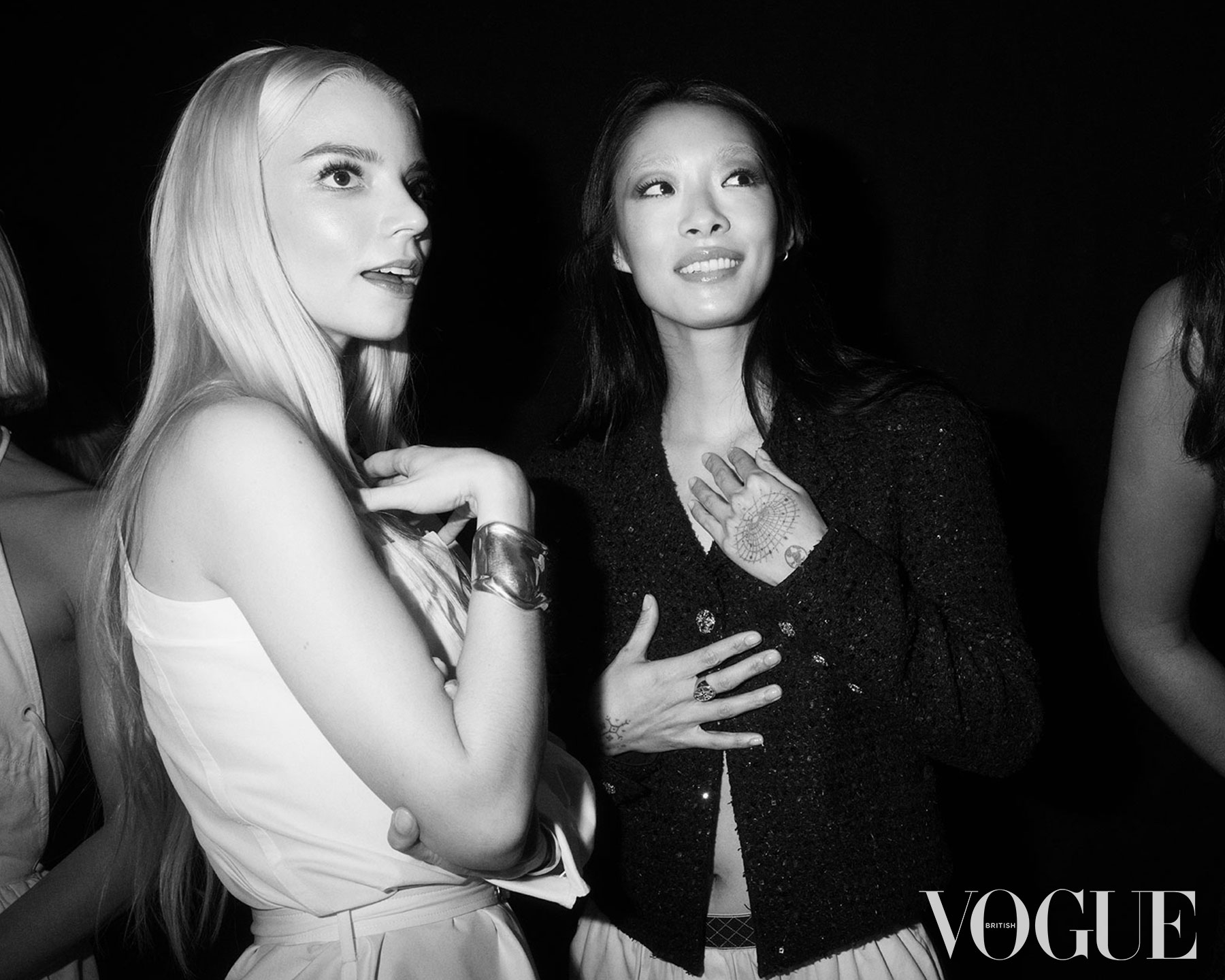 A black and white image, shows Anya Taylor-Joy, a white woman, and Rina Sawayama, a woman of Japanese heritage, apparently mid conversation. Both are looking at something off camera. Anya is holding her right hand to her chest, and Rina holds both hands in front of her chest