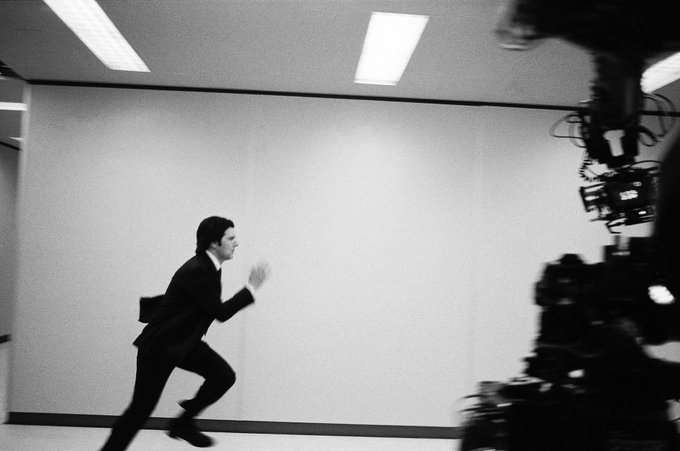 A black and white action shot of Mark S. (Adam Scott) running down a long white hallway. On the right side of the picture is a large camera recording the scene, confirming Severance Season 2 is in production.
