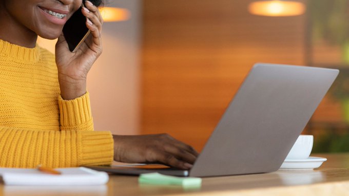 Photo of someone in a yellow sweater sitting at a laptop on the phone.