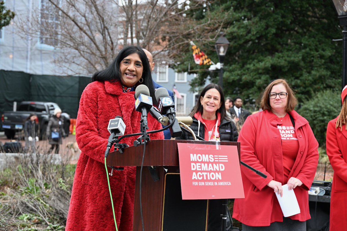 Lieutenant Governor Miller wears a red fuzzy coat as she stands at a podium that reads "Moms Demand Action for Gun Sense in America." Behind her, Moms Demand Action Advocates stand in red shirts. 