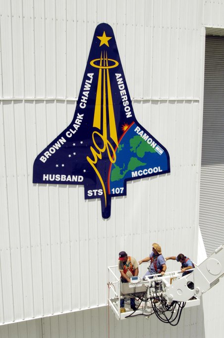 Workers lower their bucket after placing the final piece of the mission patch for STS-107 on the outside of the RLV Hangar at KSC in April 2003. The hangar was the site of the Columbia Reconstruction Project, where pieces of debris from Columbia were collected and identified as part of the mishap investigation.