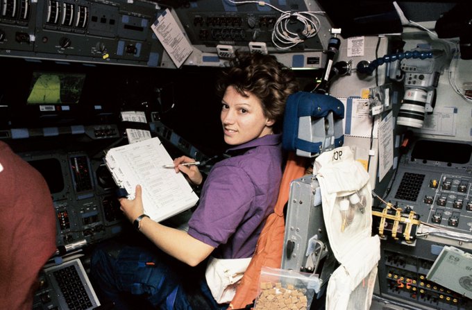 Eileen Collins sits inside the flight deck of a space shuttle. Her head is turned toward the camera as she smiles, holding a paper and pen.