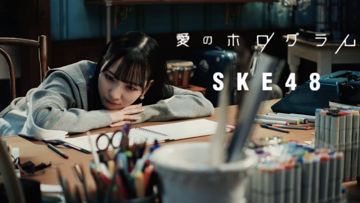 A promotional image features a young woman resting her head on her arms on a desk covered in art supplies, with the Japanese text "愛のホログラム" and "SKE48" displayed.