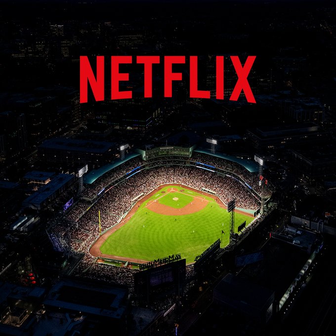 A wide aerial shot of a night game at Fenway with the city lights surrounding a brightly lit and full ballpark. A large Netflix logo is displayed above Fenway. 